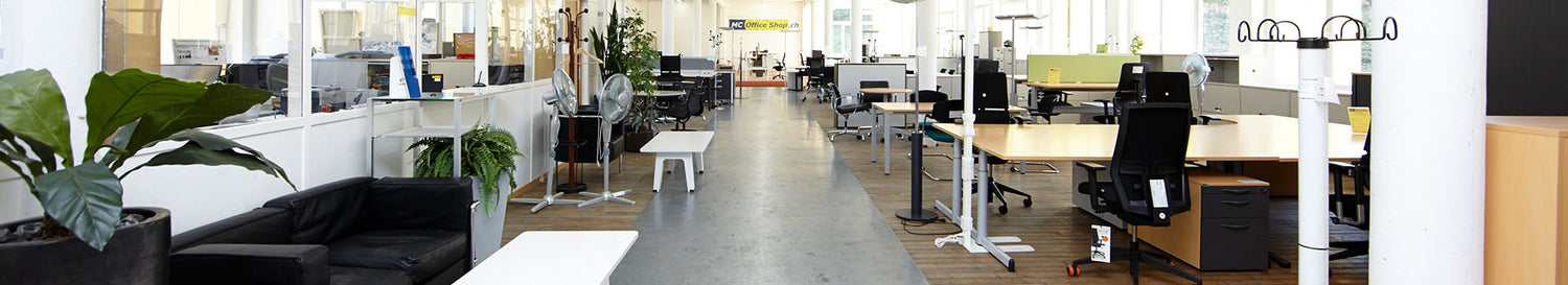 Helles Grossraumbüro mit modernen Arbeitsplätzen, Bürostühlen, schwarzem Sofa, Pflanzen und Glastrennwänden in einem offenen Showroom.