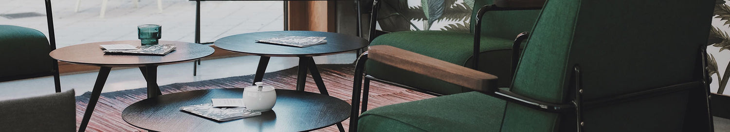 Lounge-Ecke mit grünen Sesseln und mehreren runden Couchtischen in Schwarz und Holzoptik, darauf Zeitschriften und Teegeschirr.