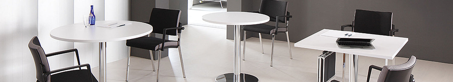 Weisser Besprechungsbereich mit runden Stehtischen und einem quadratischen Tisch, dazu schwarze Konferenzstühle auf hellem Boden.